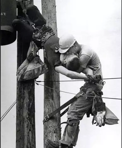 Old black and white photograph showing a lineman worker unconscious, hanging from the top of the pole where he was doing some work. A fellow worker is holding him and giving him mouth to mouth resuscitation.