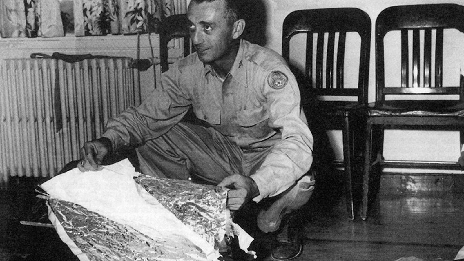 Army officer kneeling on the floor holding pieces of foil and other debris.