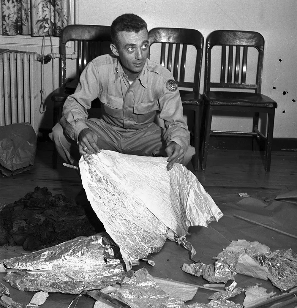 Army officer kneeling on the floor holding pieces of foil and other debris.