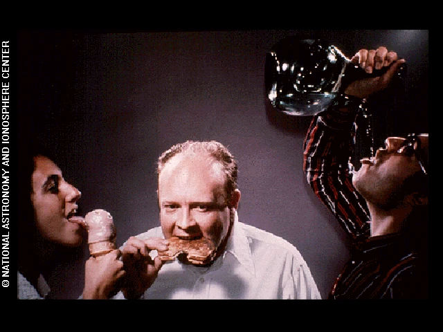 Photo shows a man eating something fried or crispy. There is another man next to him drinking water from a jug, and there is a woman in the photo licking ice-cream.