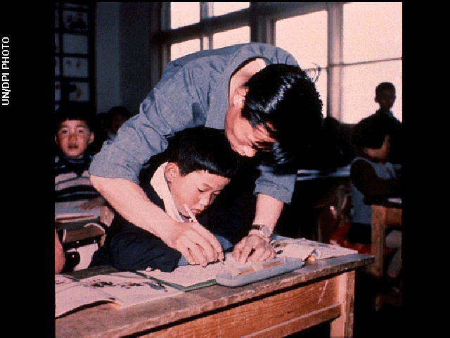 Photo shows a teacher helping explain something to a boy learning in school.