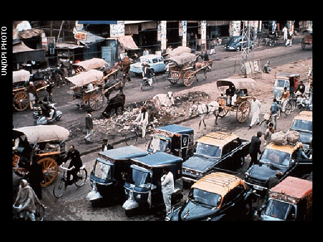 Photo shows a busy street somewhere in Asia, perhaps India or Thailand. There are many motorbikes, cars, horses with carriages, bicycles and tuk-tuk vehicles.