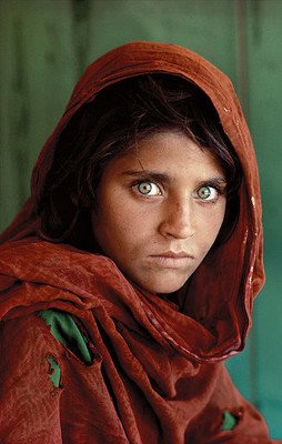 Photograph of an Aghan girl. She looks scared and shocked, looking straight at the camera. She has a look as if she has never seen a camera before. Photo has a bright colorful contrast to it which makes it unique and appealing. She is wearing a dark red headscarf, and the background is dark green, she also has pastel green bright eyes, and she has rips in her shroud on the arm and underneath the rips you can see a dark green undergarment.